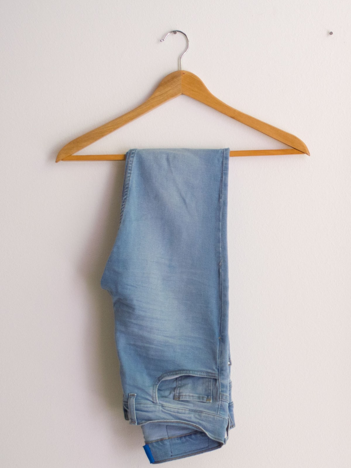 Lichtblauwe gewassen damesjeans over een houten kleerhanger tegen een effen witte muur
